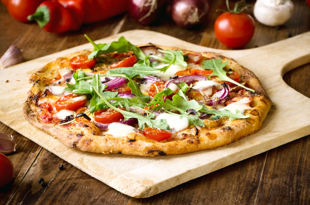 This photo shows a pizza, placed on a wooden peel, that's topped with arugula, tomatoes, red onions and mozzarella. It's surrounded by other veggies like tomatoes, mushrooms and red peppers.