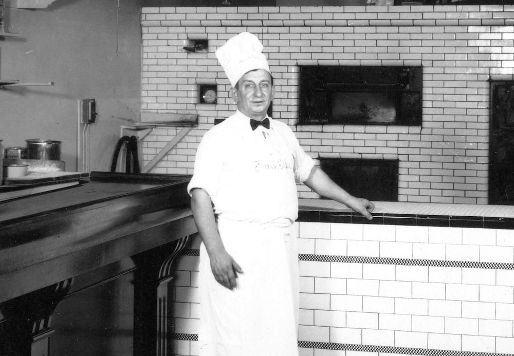 This vintage black-and-white photo shows Frank Pepe, clad in a white chef hat, apron and shirt, in his kitchen at Frank Pepe Pizza Napoletana