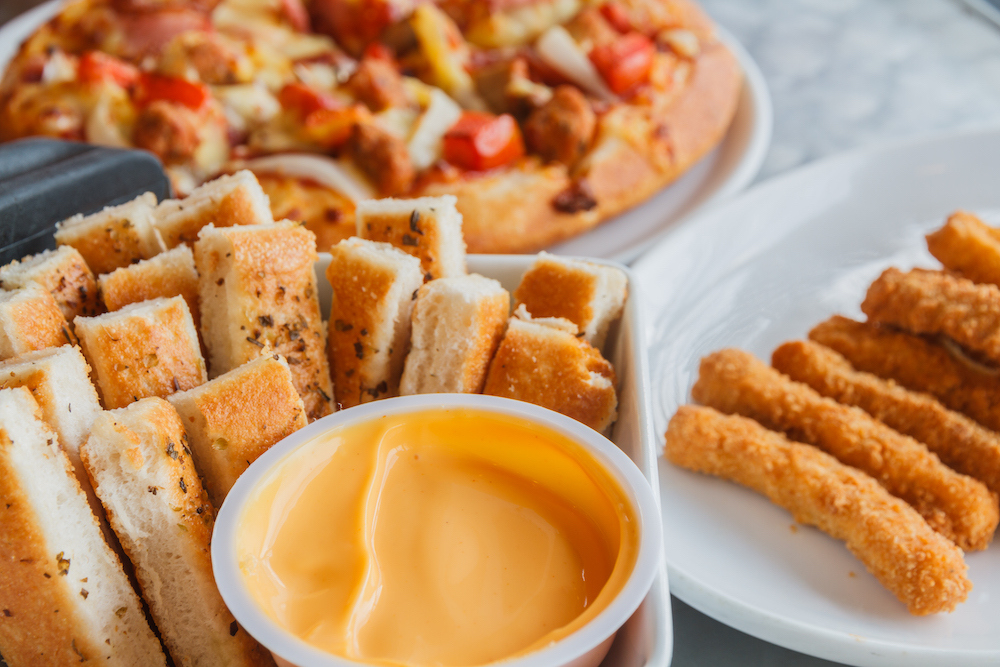 Delicious-looking breadsticks with a dipping sauce placed next to a pizza and fried food