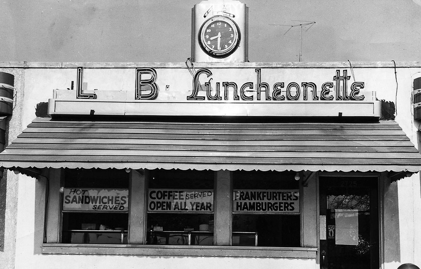 This vintage black-and-white photo shows the exterior of one of the L&B Spumoni buildings with a sign in front that reads "L&B Luncheonette."