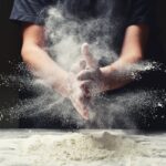 This photo shows a man clapping his hands together and scattering flour while making pizza dough in a restaurant kitchen.