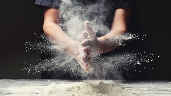 This photo shows a man clapping his hands together and scattering flour while making pizza dough in a restaurant kitchen.
