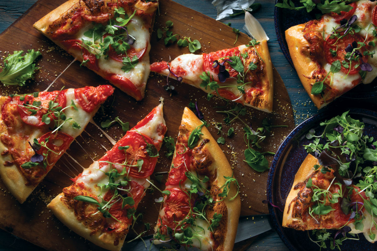 This photo shows artfully arranged slices of pizza topped with tomatoes, lots of mozzarella and arugula on a wooden peel, with additional slices nearby on dark-blue or black plates.
