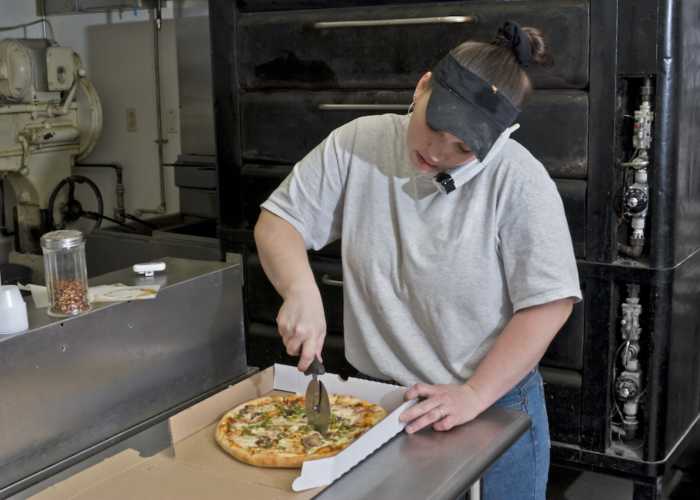 the photo shows a pizzeria employee practicing phone etiquette while slicing a pizza