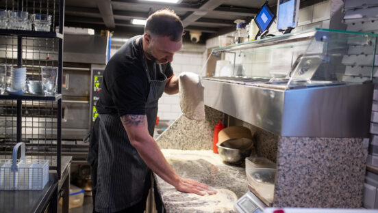 This photo shows a Pizzaiolo stretching dough in his hands for pizza and spreading flour on the kitchen counter.