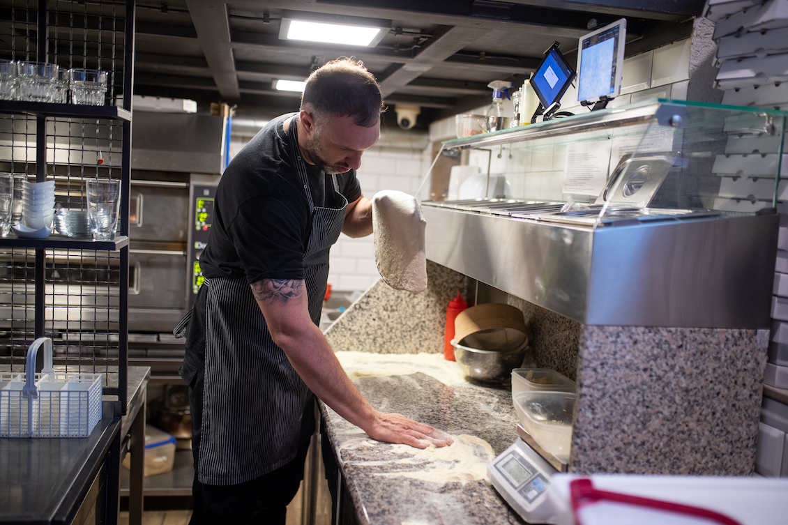 This photo shows a Pizzaiolo stretching dough in his hands for pizza and spreading flour on the kitchen counter.
