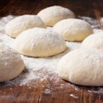 This photo shows a batch of rolled dough balls placed on a wooden board, with a lot of flour sprinkled throughout.