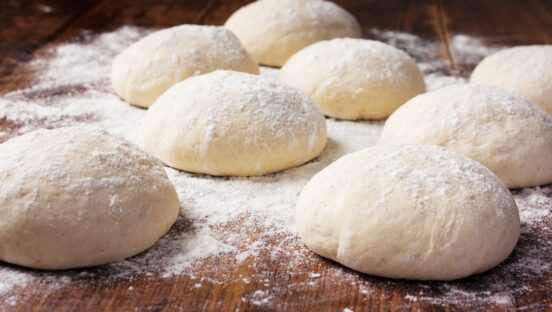This photo shows a batch of rolled dough balls placed on a wooden board, with a lot of flour sprinkled throughout.