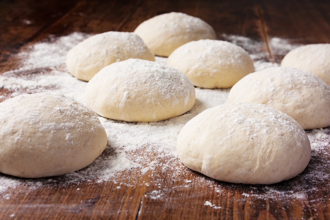 This photo shows a batch of rolled dough balls placed on a wooden board, with a lot of flour sprinkled throughout.