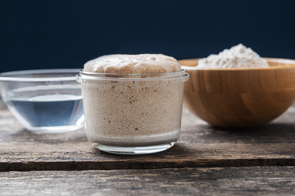 This photo shows a heaping jar of sourdough starter yeast with a cup of flour and water next to it, all on a rustic wooden desk.