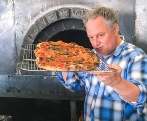 Chris Bianco holds a baked pizza on a screen close to his face and peers one it at the camera.