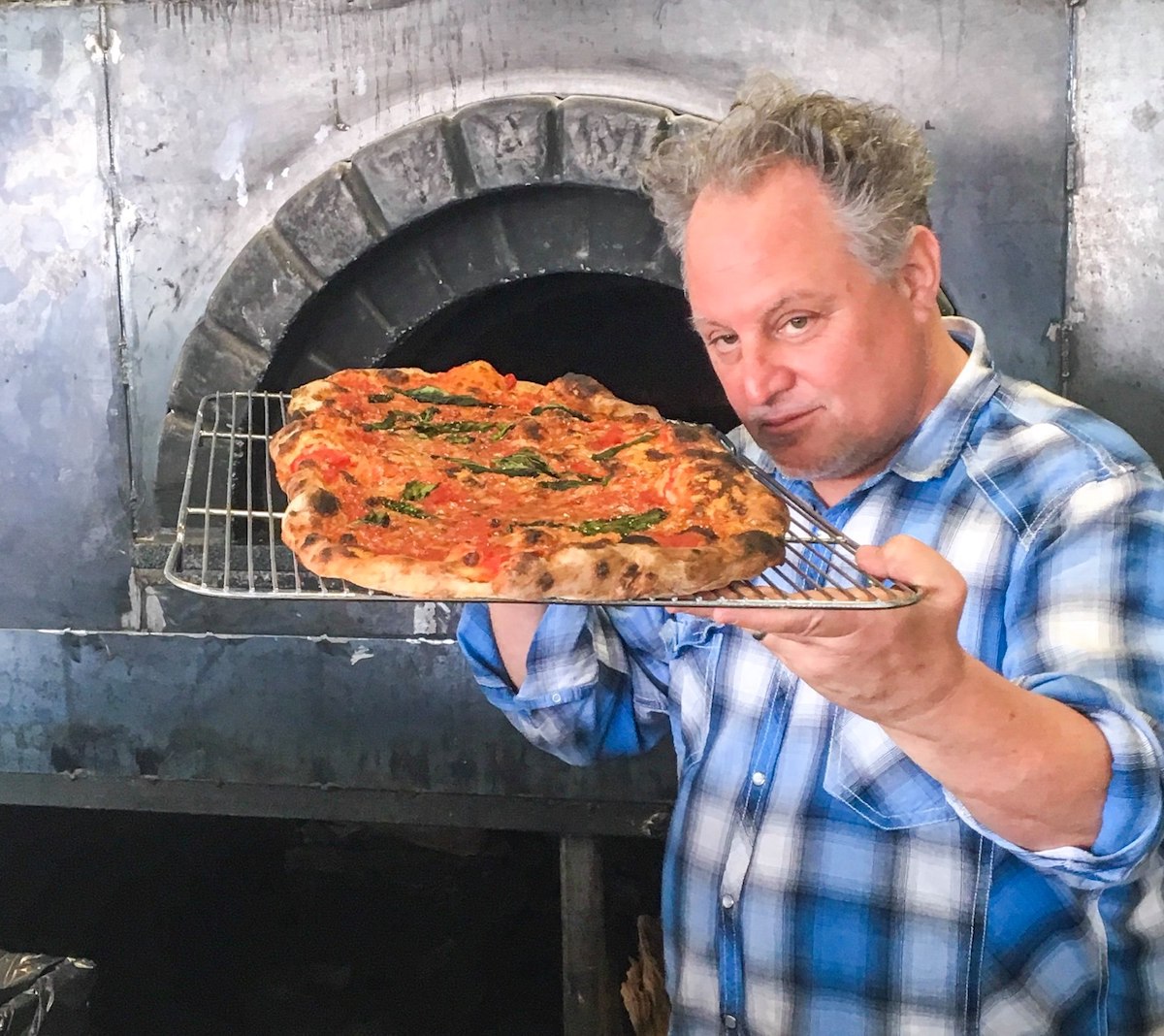 Chris Bianco holds a baked pizza on a screen close to his face and peers one it at the camera.