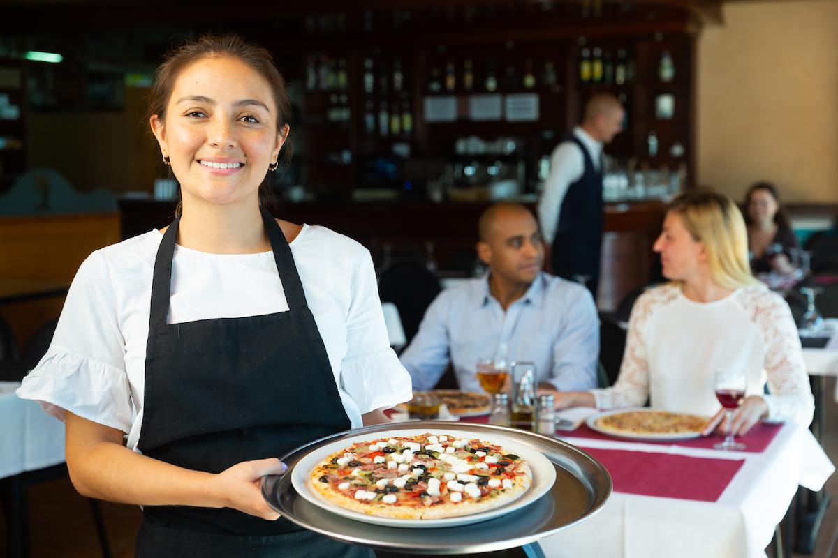 a smiling woman with a pizza in a tray stands in front of a table of customers enjoying their food