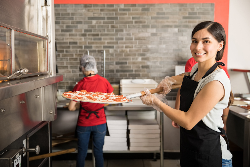 Smiling chef wearing uniform putting raw pizza in modern oven for baking while looking at camera and staff working in background in pizza shop