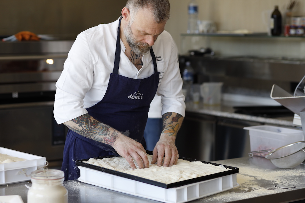 this photo shows Roman pizzaiolo Gabriele Bonci in a white shirt and dark blue apron pressing out dough in a tray for a Roman pizza at Pizzarium in Rome, Italy.