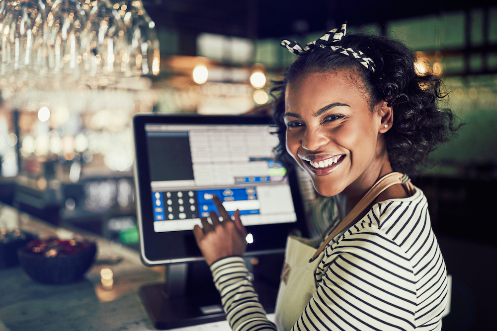 an attractive black woman in a striped shirt smiles as she keys in information in a restaurant's POS system