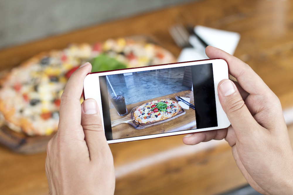 A man's hand shoots a photo of a colorful pizza with a mobile phone, close up