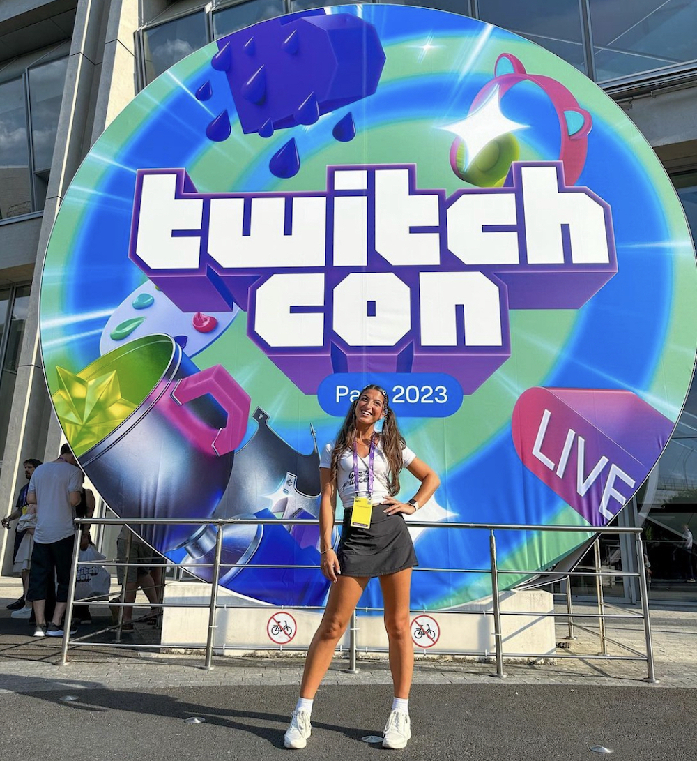 This photo shows Giuliana, wearing a short black skirt and white t-shirt, posing in front of the TwitchCon sign in Paris, France.