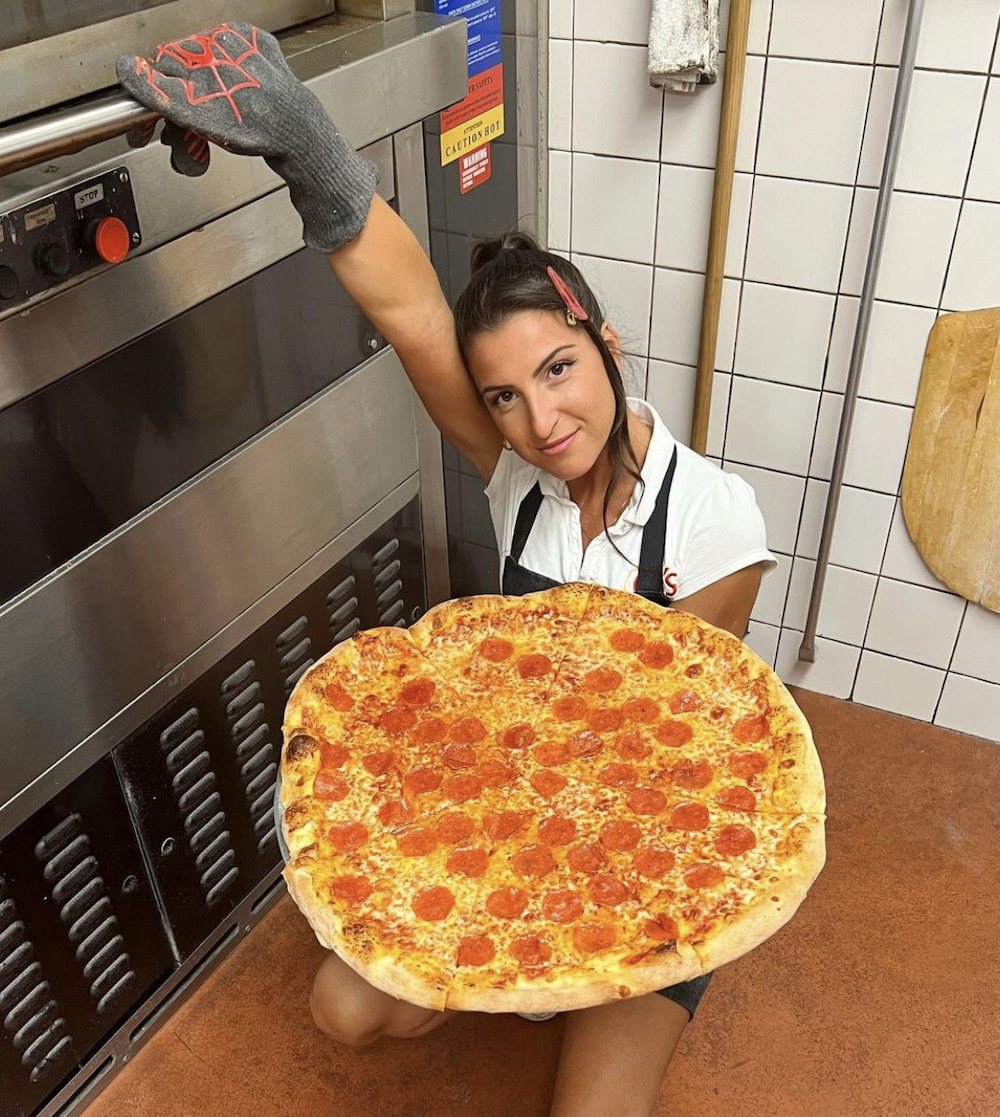 Giuliana kneels down beside her pizza oven and peers up at the camera, holding a huge pepperoni pizza