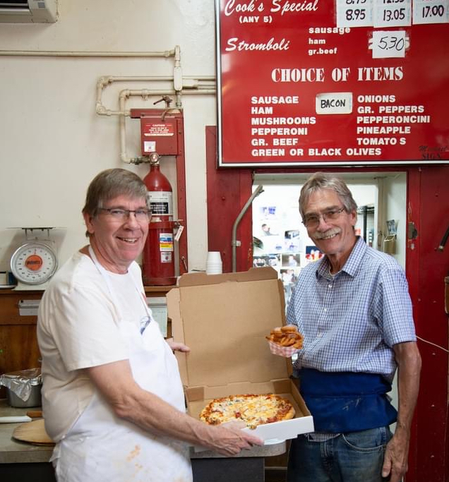 the Cook brothers stand in front of a large red menu board and hold up a box with a pizza
