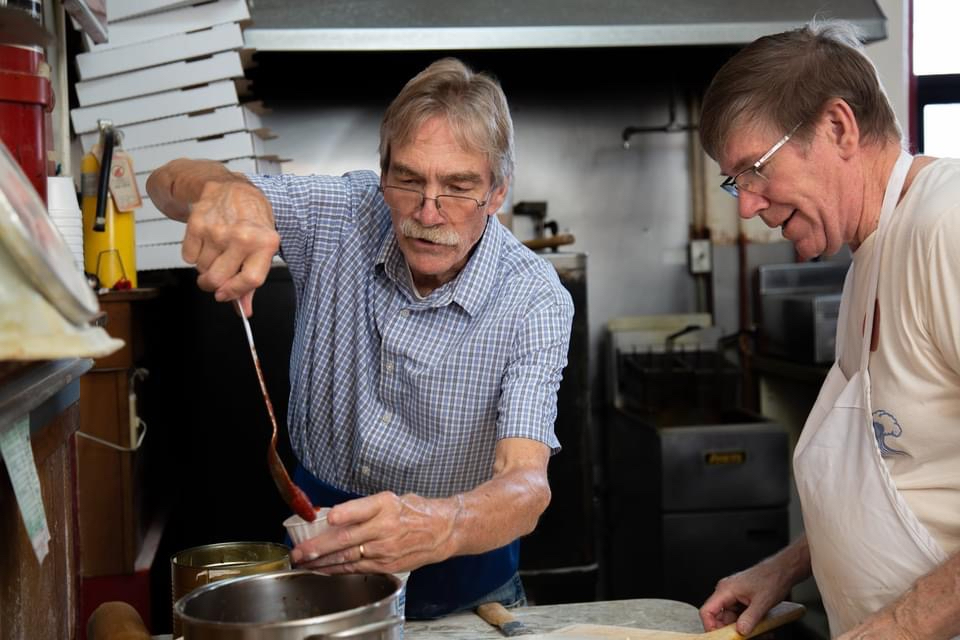 this photo shows Stan and Steve Cook using a metal ladle to pour some marinara sauce into a dipping cup in the kitchen of Cook's Pizza