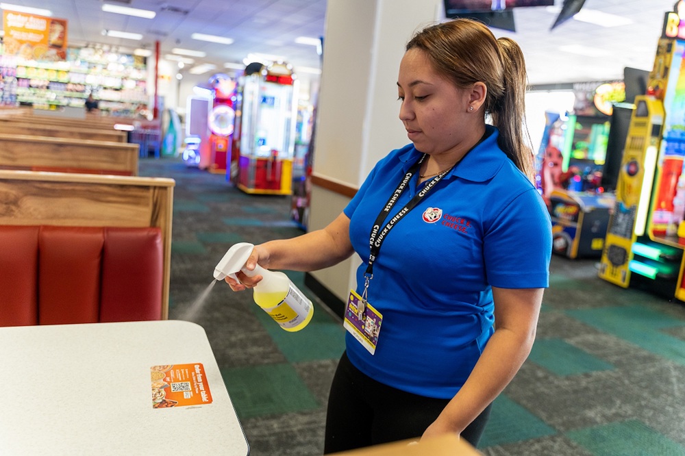 a woman in a blue Chuck E. Cheese uniform shirt sprays down a table and gets ready to wipe it.