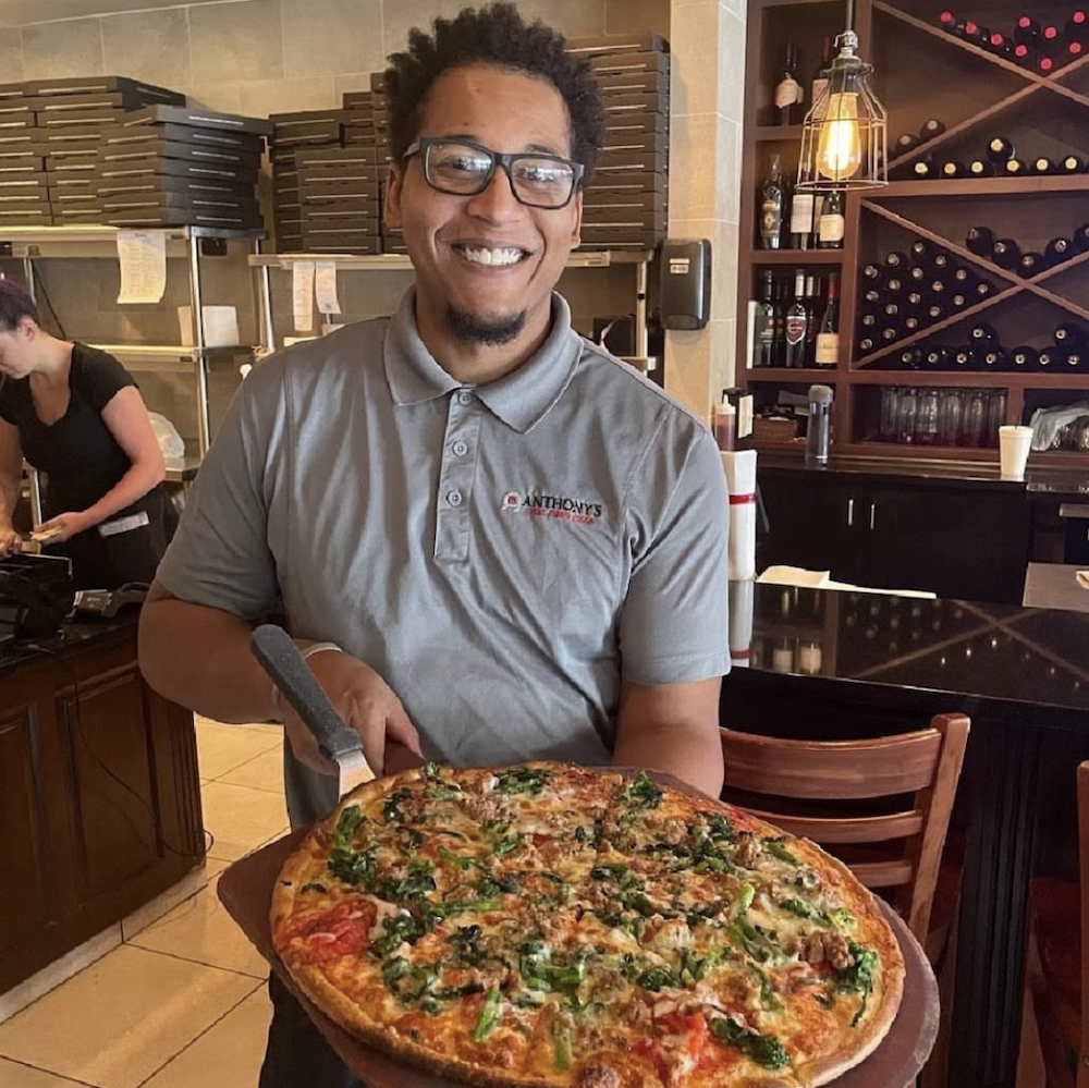 a smiling, bespectacled African-American man in a light blue shirt holds out a large pizza with a wide variety of toppings