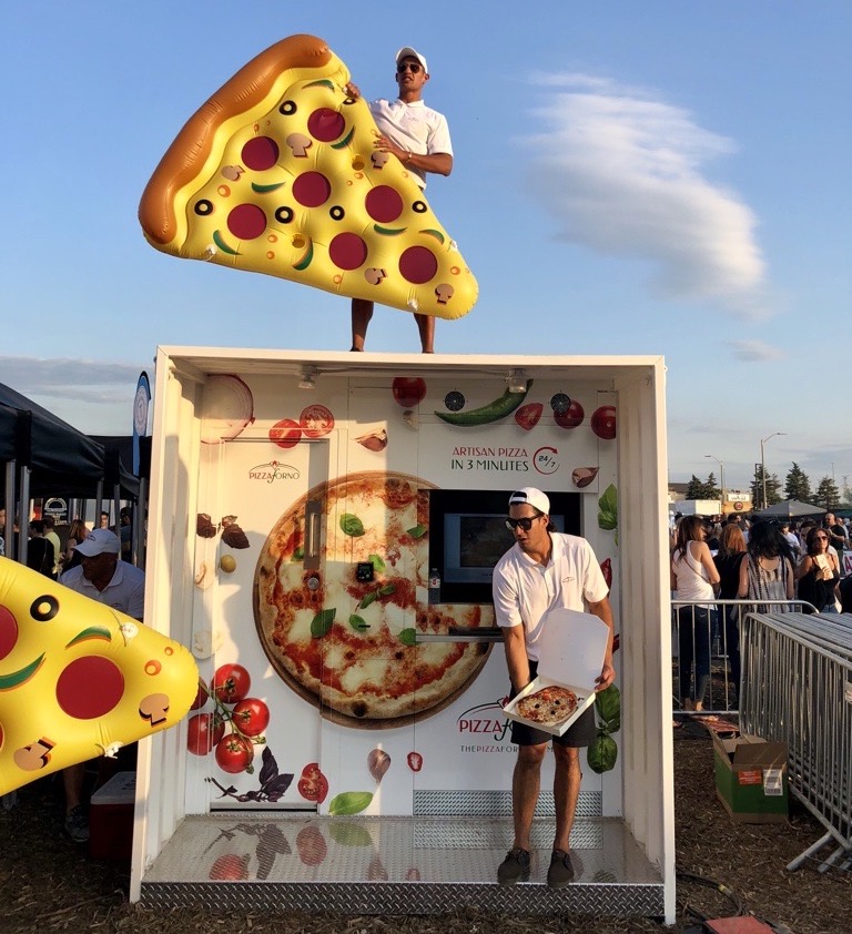 a man in a white shirt and cap stands in an enclosed PizzaForno kiosk, holding a boxed pizza, while another man stands on top of the kiosk with an inflatable red, yellow and orange pizza slice