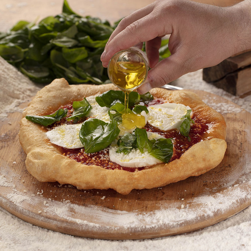 in this photo, a bottle of olive oil is poured onto a beautiful Margherita pizza