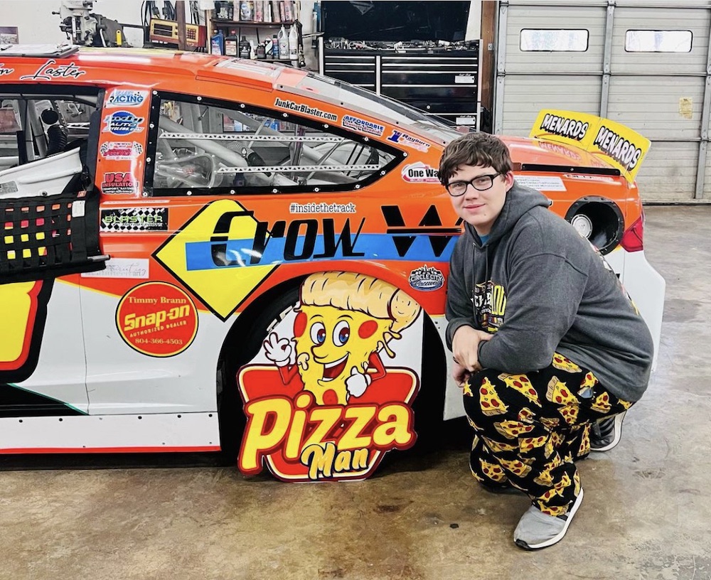 Brayton Laster, wearing pepperoni pizza-themed pants, kneels next to a race car with a Pizza Man Pit Road sign
