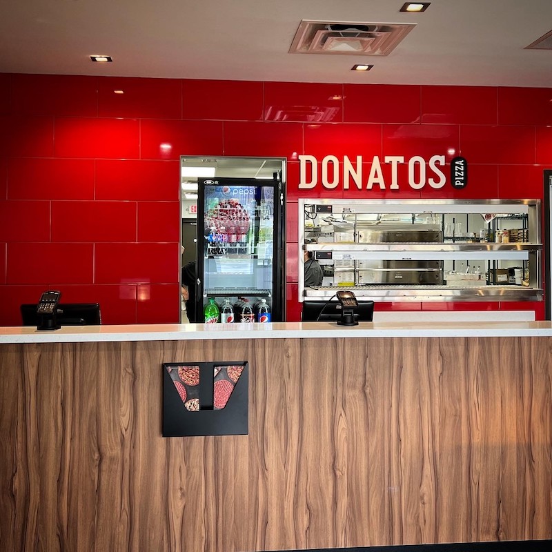 the interior of a Donatos restaurant with a red, tiled wall in the background and a white-lettered sign that reads Donatos just above an entrance to the pizza kitchen