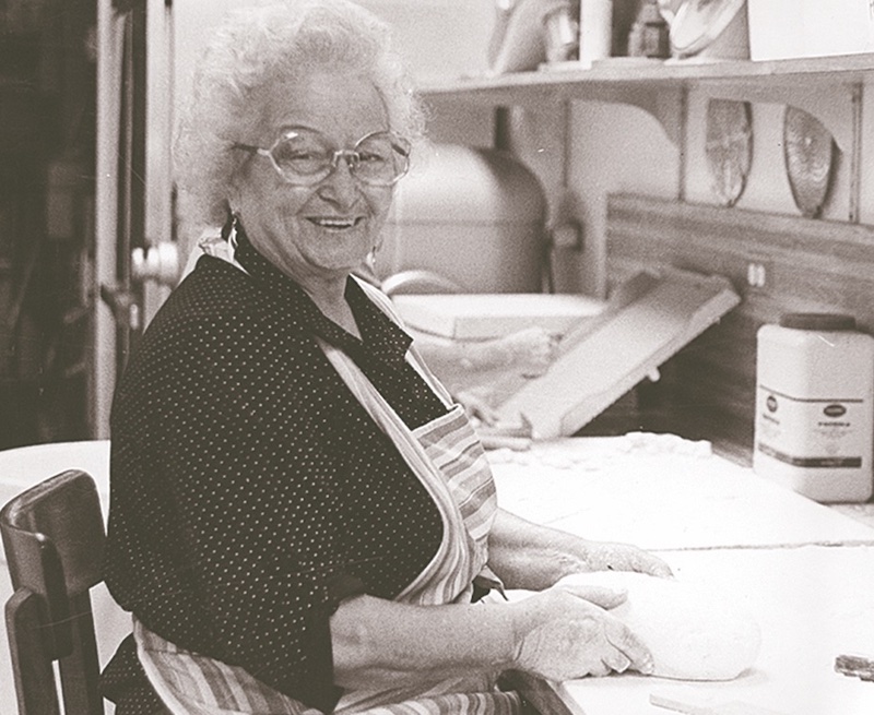 this black-and-white photo shows a smiling Mary Fazio with gray hair as she works with a ball of pizza dough in her kitchen