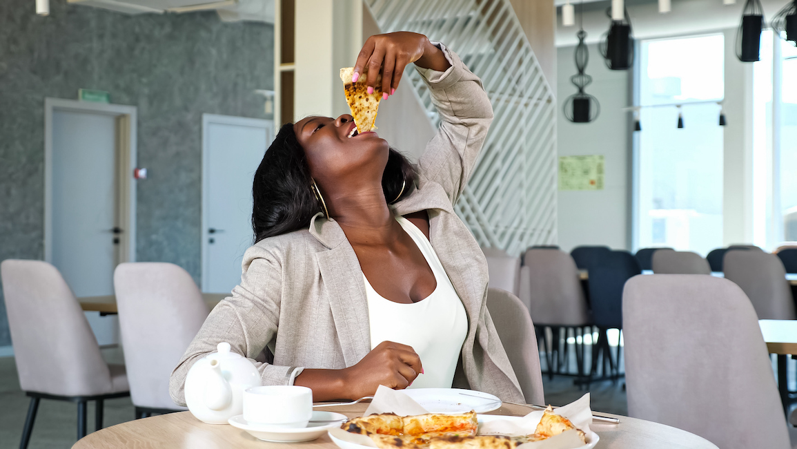 Positive African-American woman in grey jacket enjoys eating delicious pizza at table with teapot and cup in light contemporary cafe