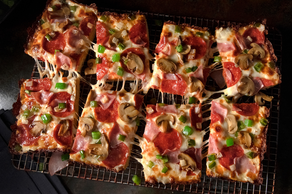 this photo shows eight slices of a Detroit-style pizza topped with pepperoni, mushrooms, ham, onions, and bell peppers, set against a black background