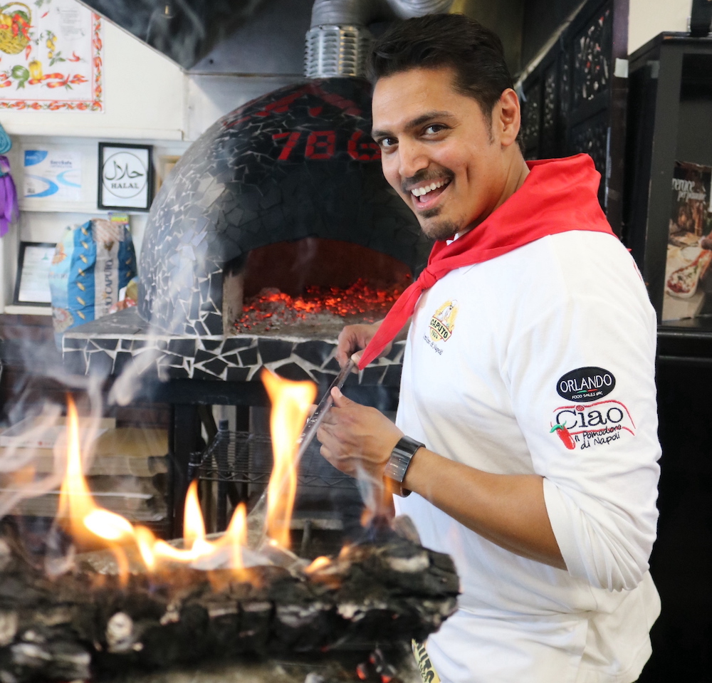 Ali Haider, wearing a white chef's coat with a red scarf, stokes a fire in his pizza kitchen while smiling at the camera