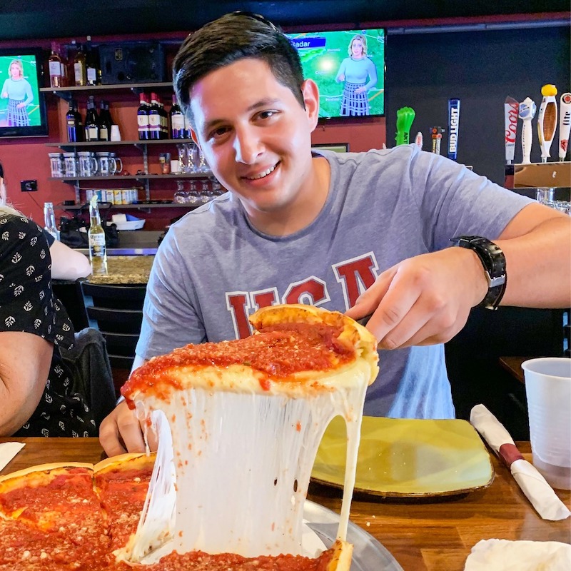 a happy-looking young man in a t-shirt takes a slice of deep-dish pizza with a long cheese pull at Angie's Sports Bar and Pizzeria