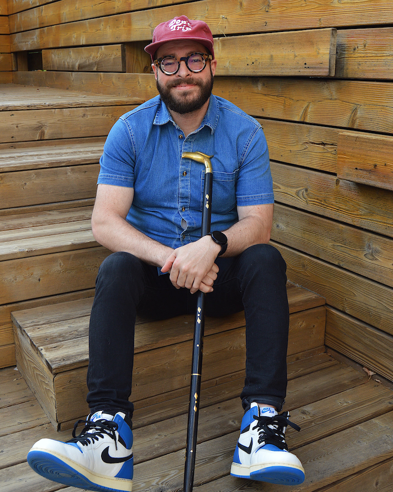 Here we see Billy sitting on a set of steps with his cane in his hand. He's wearing a blue denim shirt and a maroon cap 