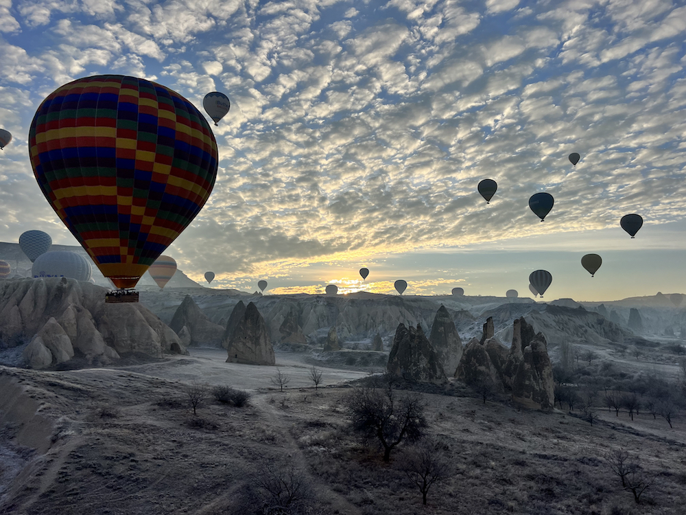 this photo shows huge hot air balloons floating over the fantastical rock formations, called fairy chimneys, in Goreme, Turkey, as the morning sun peeks over the horizon