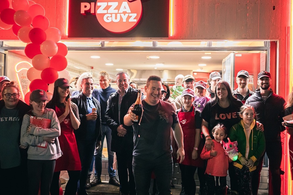 Ciaran Kelly, wearing a black t-shirt and pants and short hair, has just popped a bottle of champagne while surrounded by employees and customers at the grand opening of a new location.