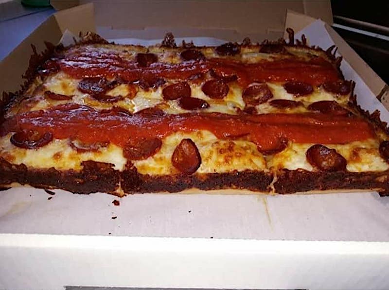 this is a shot of a Detroit-style pizza, shown in a carryout box, with stripes of tomato sauce on top of the cheese and crispy edges