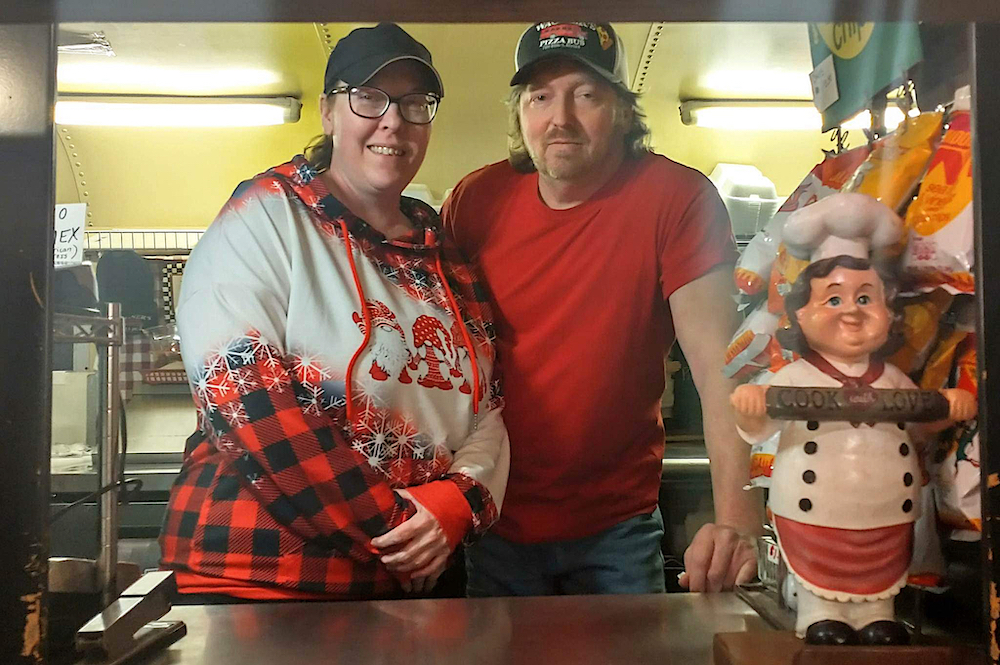 this photo shows Diana and Mike Wagner peering out of the ordering window of Wagner's Pizza Bus with a small statue of a man with a chef's hat and a rolling pin