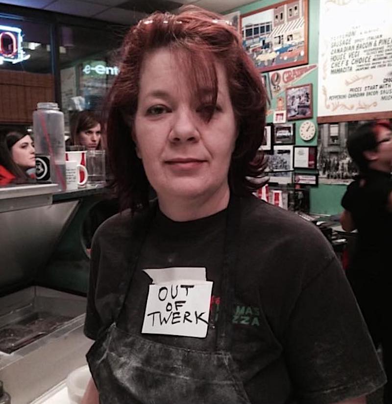 the photo shows Krista Nelson, with short reddish hair and wearing a black t-shirt with an attached note that reads Out of Twerk