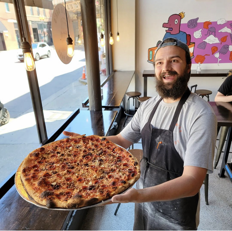 a smiling man, wearing a backwards cap and an apron, shows off a whole pizza topped with pepperoni and sausage