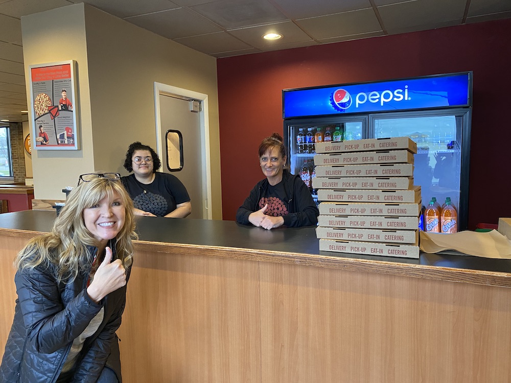 Jane Grote Abell is seen to the left, giving the thumbs up and smiling while two employees stand at the ordering counter with a stack of pizza boxes on the right-hand side