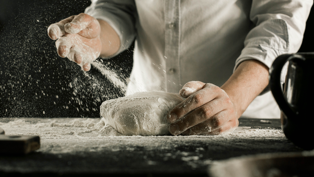 Male chef hands knead dough with flour on kitchen table, shown from a side view