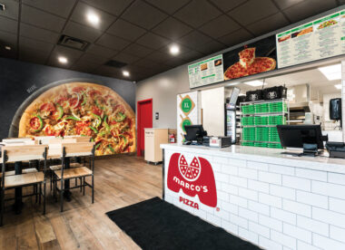 the interior of a Marco's Pizza restaurant. in the foreground is a white ordering counter with the red Marco's logo, and in the background is a large mural depicting a pizza