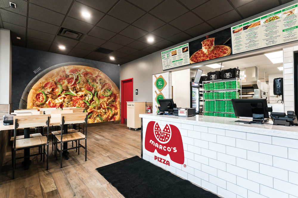 the interior of a Marco's Pizza restaurant. in the foreground is a white ordering counter with the red Marco's logo, and in the background is a large mural depicting a pizza