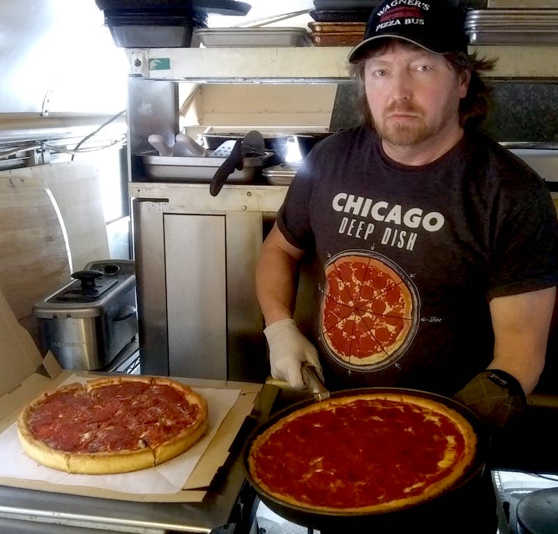 Mike Wagner, wearing a black Chicago Deep-Dish t-shirt and Wagner's Pizza Bus cap, displays a pair of Chicago deep-dish pizzas