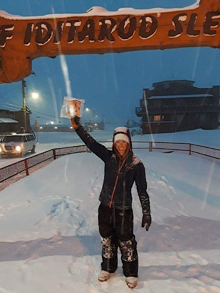 a woman in a sledding uniform holds up a slice of pizza in a zip-lock bag while standing in front of the finishing line of the Iditarod Dog Sledding Race.