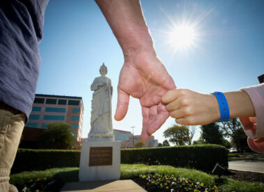 close-up of a man's hand and a child's hand (with a hospital bracelet) in front of a statue of St. Jude on the St. Jude Children's Research Hospital campus